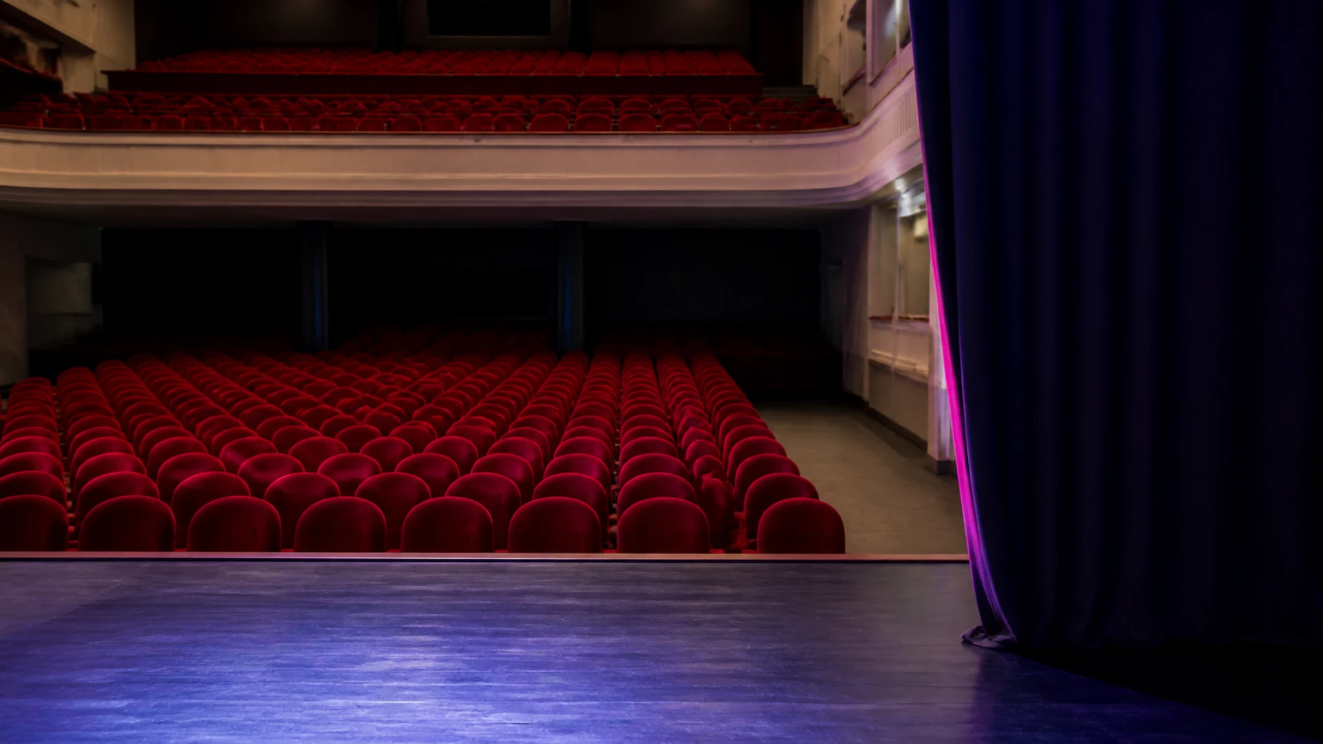 Salle de spectacle vue depuis la scène avec fauteuils vides et rideau partiellement ouvert