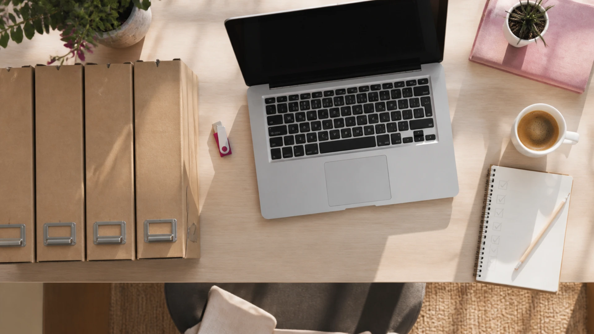 Vue de dessus d’un bureau clair avec quatre boîtes d’archives en carton, une clé USB rose, un ordinateur portable ouvert, une tasse de café et un carnet de cases cochées