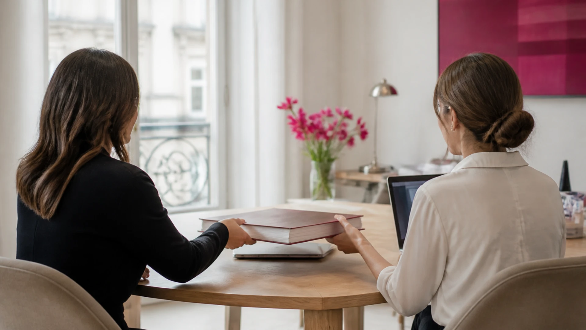Deux femmes vues de dos à une table en chêne, l’une fait glisser un épais registre relié bordeaux vers l’autre, devant une fenêtre haussmannienne et un ordinateur portable ouvert