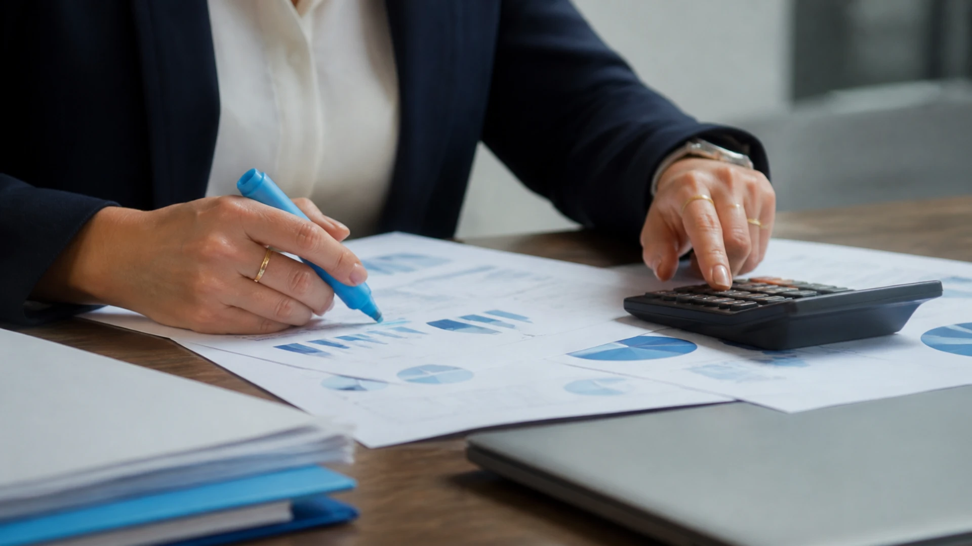 Mains d’une professionnelle qui surligne des graphiques imprimés et tape sur une calculatrice, à côté d’un ordinateur portable fermé et d’une pile de dossiers